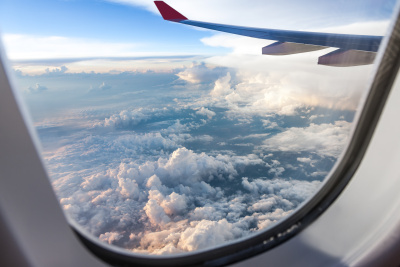 Aufnahme aus einem Flugzeugfenster mit Blick auf einen Flugzeugflügel und auf den blauen Himmel mit Wolken.