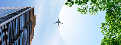 Key Visual Startseite Zwei Bilder werden durch einen geschwungenen Bogen, dem CENA-Bogen, voneinander optisch getrennt. Auf dem linken Bild fliegt ein Flugzeug am Himmel über eine Stadtlandschaft. Am linken Bildrand ragt ein gläsernes Hochhaus in den Himmel, während rechts grüne Baumkronen den Blick einrahmen. Der Himmel ist klar und blau, wodurch das Flugzeug deutlich sichtbar ist.